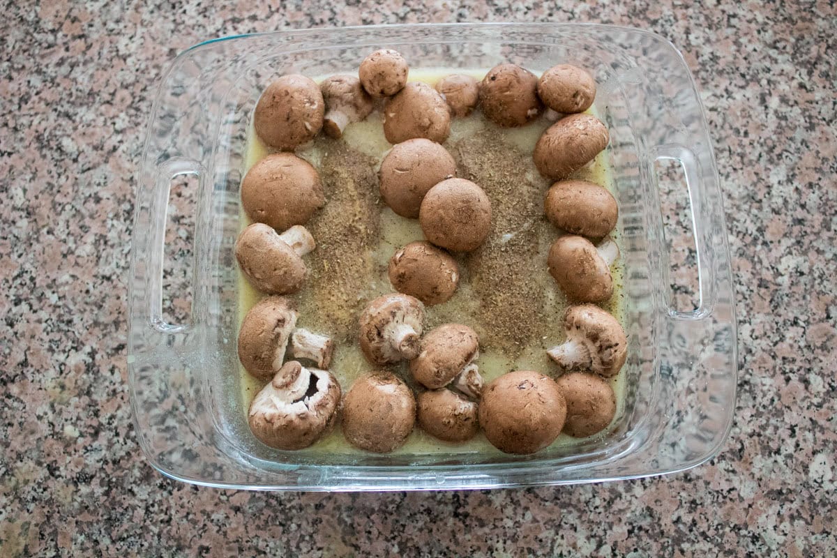 A glass baking dish filled with whole brown mushrooms arranged along the edges, sitting in a pale liquid with seasoning, on a granite countertop.