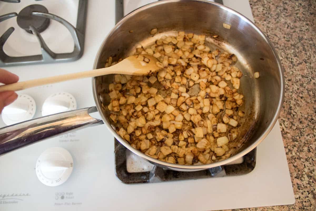 A person stirs diced onions that are browning in a stainless steel saucepan on a white gas stove with a wooden spoon.