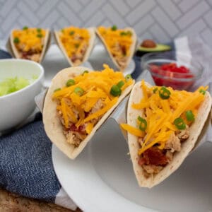 Three soft tacos filled with shredded chicken, bacon, cheddar cheese, and chopped green onions are served on a white plate, with bowls of lettuce and diced tomatoes in the background.