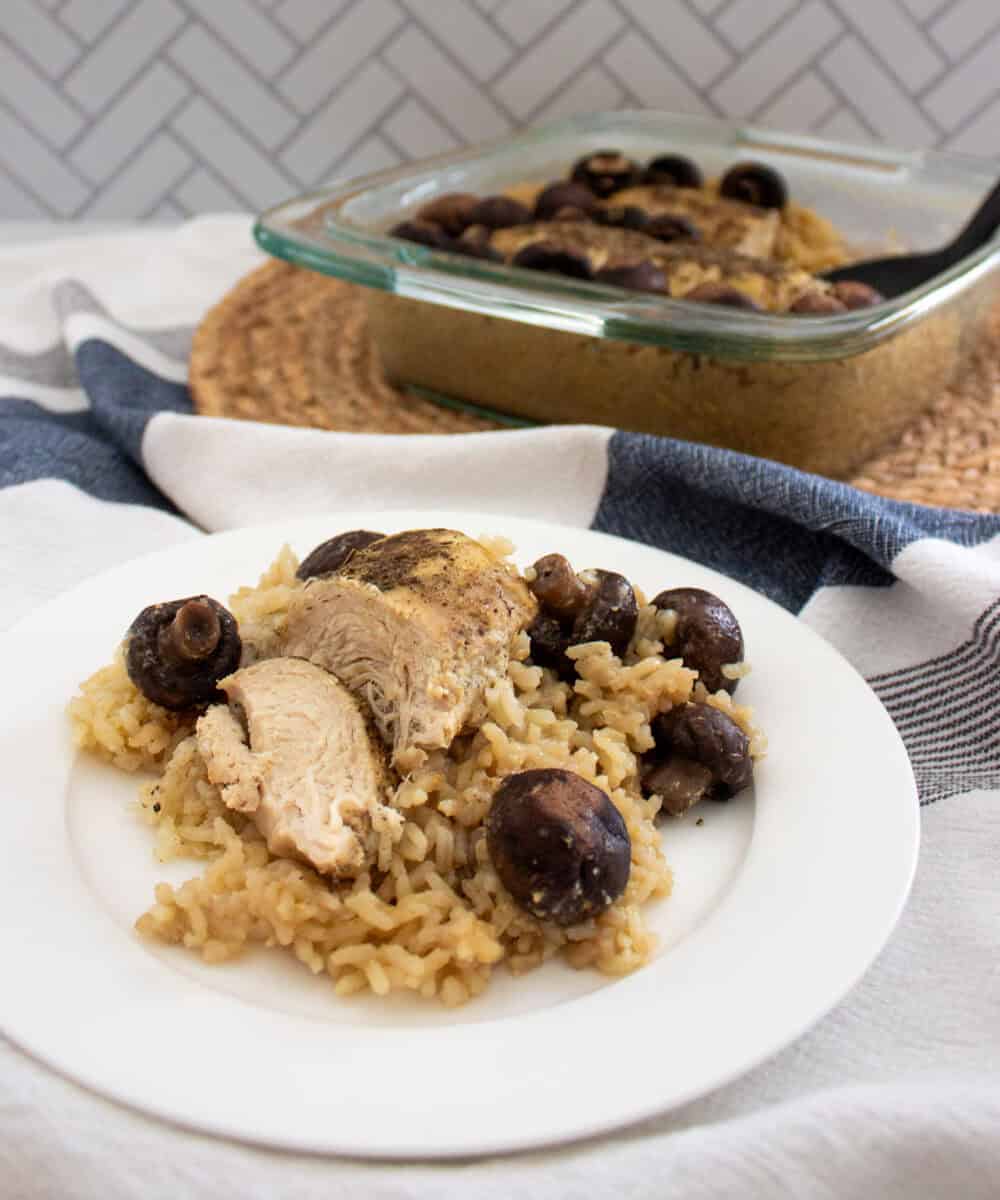 A plate with baked chicken breast, brown rice, and mushrooms sits in the foreground, while a glass baking dish with more of the same food is in the background on a woven mat. The setting includes a striped cloth.