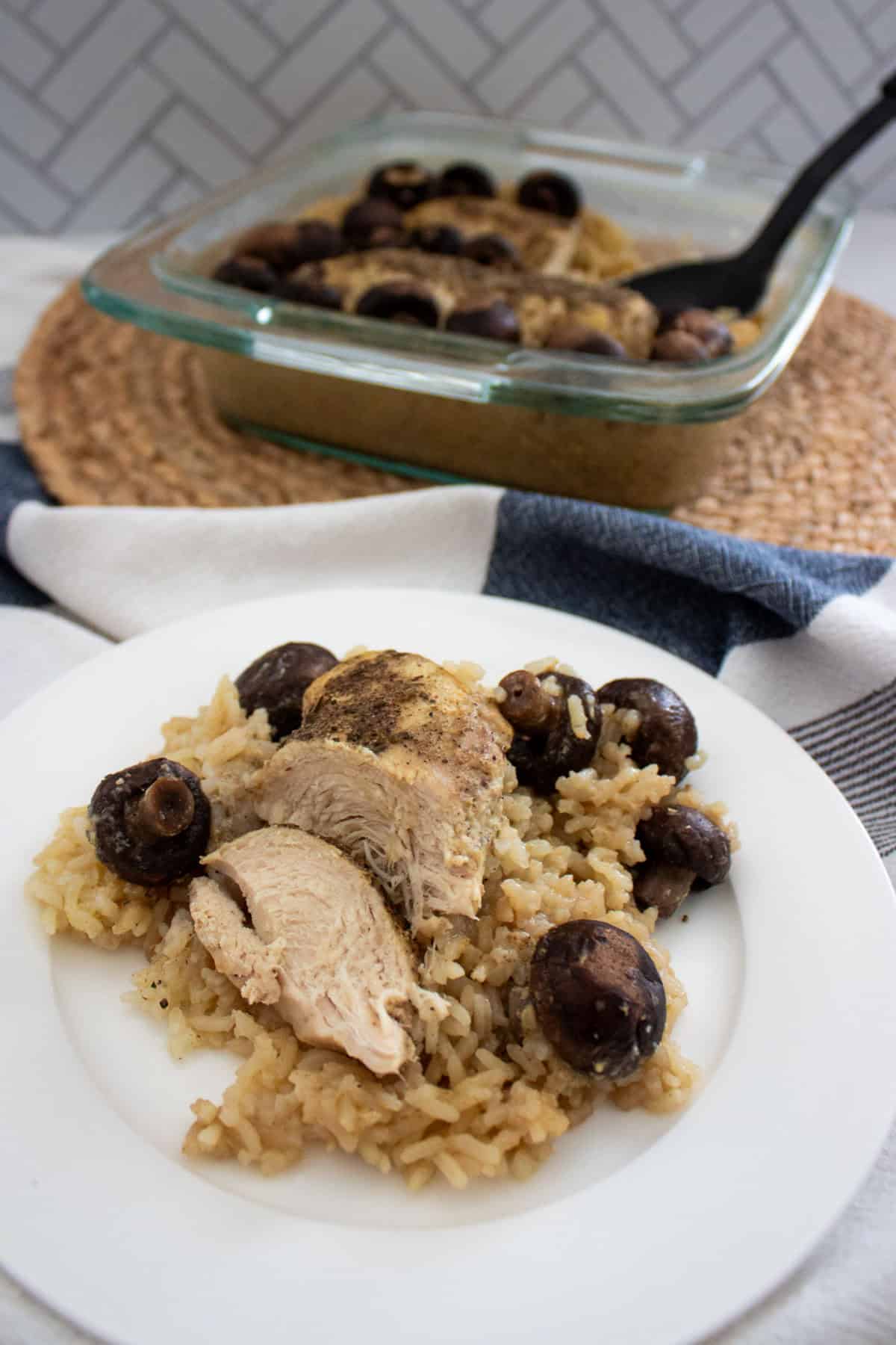 A white plate with baked chicken breast, rice, and mushrooms sits in front of a glass baking dish containing more of the same meal. A black serving spoon rests in the dish, which is on a woven mat.