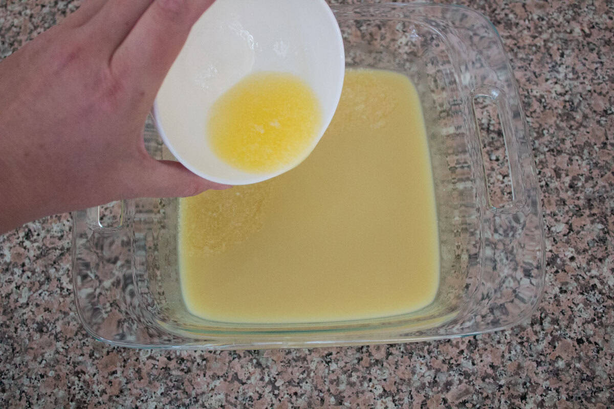 A hand pours melted butter from a white bowl into a glass baking dish filled with yellow liquid, on a speckled countertop.