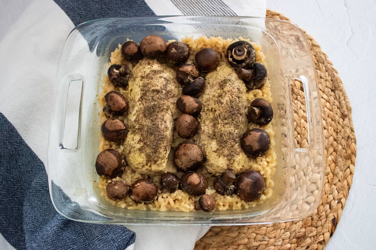 A glass baking dish with two seasoned chicken breasts, whole mushrooms, and rice, placed on a woven placemat next to a folded kitchen towel.