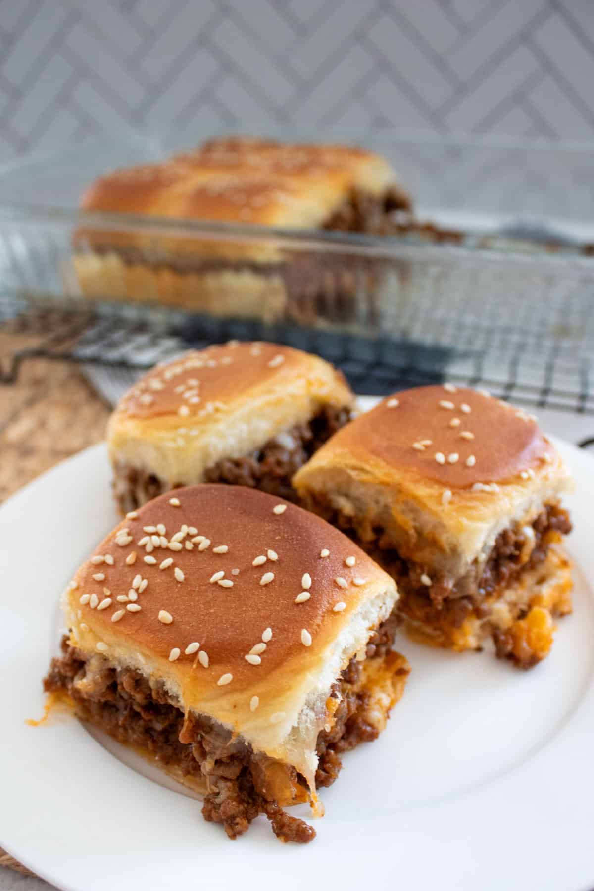 Three sesame seed-topped slider sandwiches filled with ground beef and melted cheese sit on a white plate, with more sliders in a glass baking dish in the background.