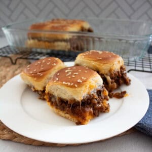 Three cheesy sloppy joe sliders with a golden top sit on a white plate. A glass baking dish with more sliders is in the background, and the filling appears to be ground beef and sauce.