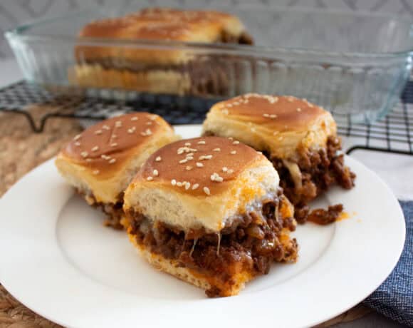 Three cheesy sloppy joe sliders with a golden top sit on a white plate. A glass baking dish with more sliders is in the background, and the filling appears to be ground beef and sauce.