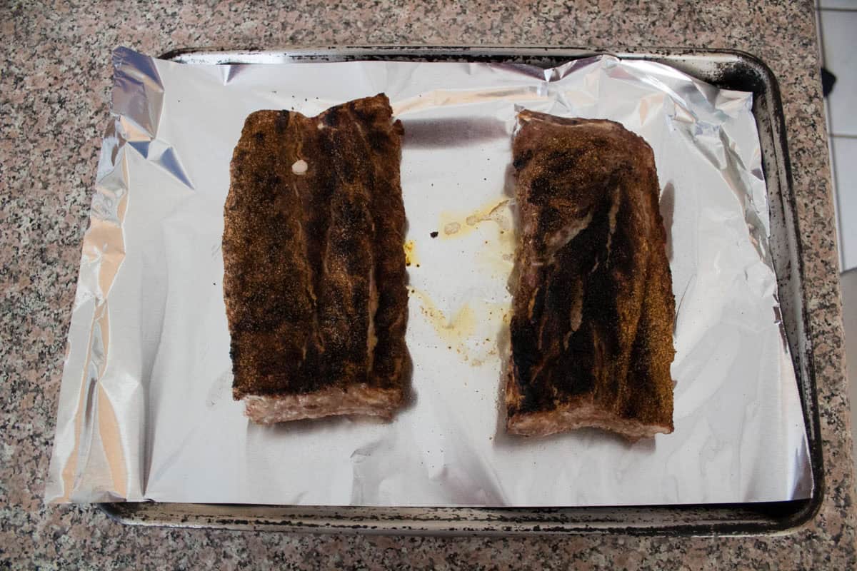 Two cooked racks of ribs with seasoning are placed on a foil-lined baking sheet, which rests on a speckled countertop. The ribs are separated, with a small gap between them.