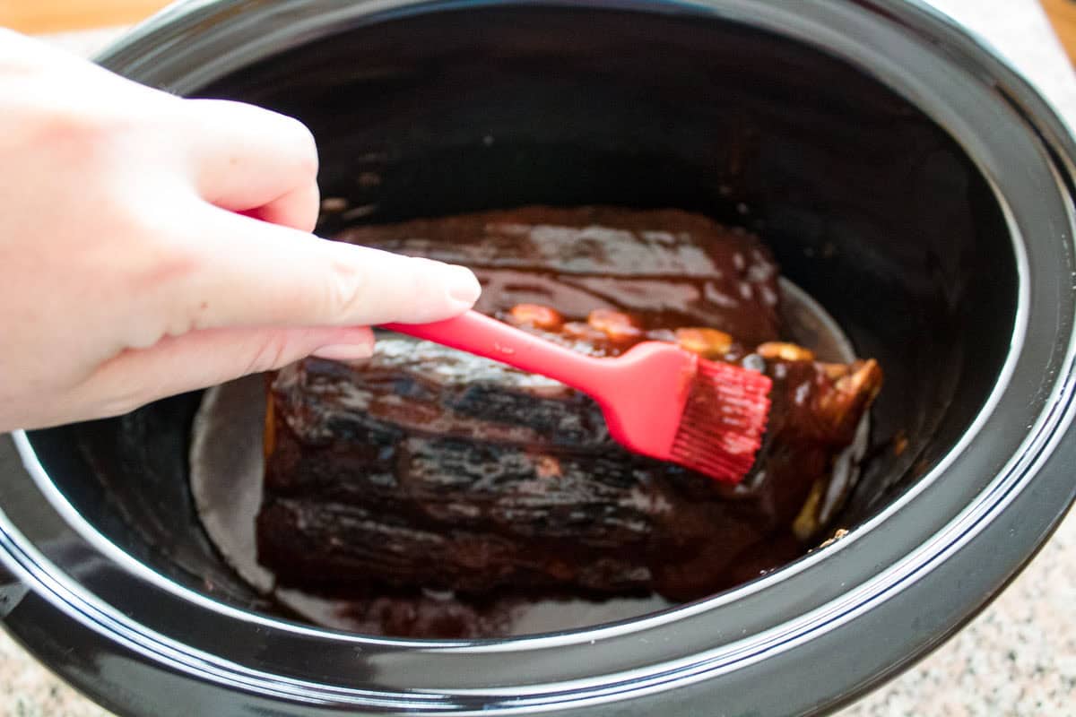 A hand uses a red silicone brush to apply sauce to a piece of meat inside a black slow cooker.