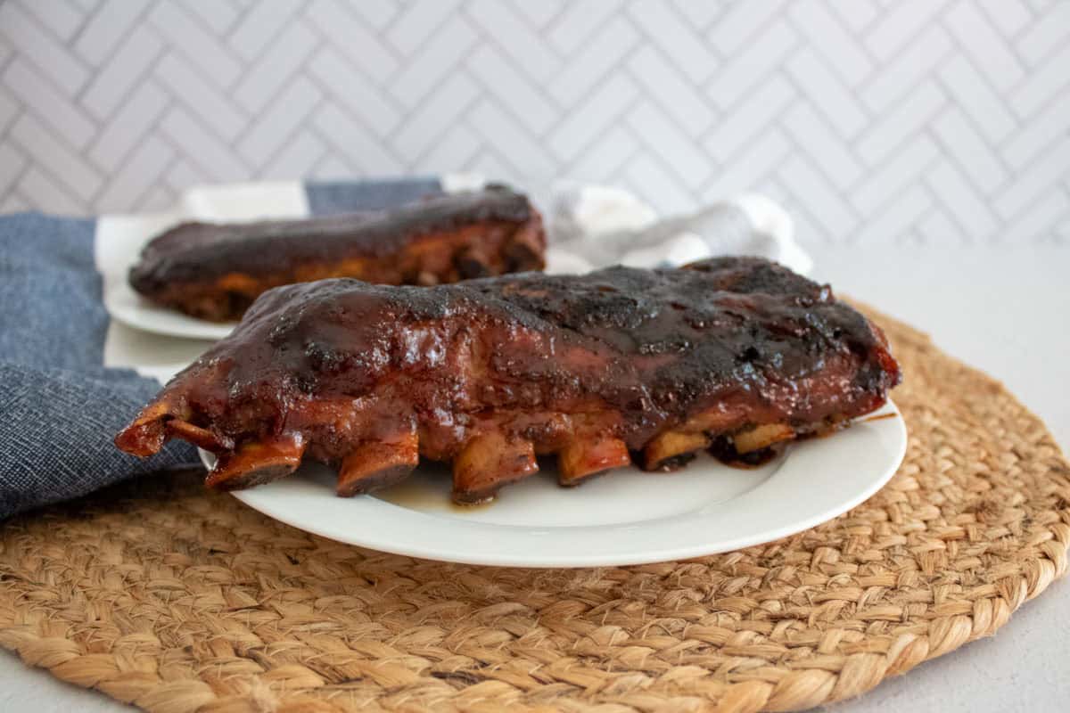 Two plates with glazed, cooked pork ribs sit on a woven placemat. The ribs are coated in a shiny, dark barbecue sauce, and a blue napkin is partially visible beside one plate.