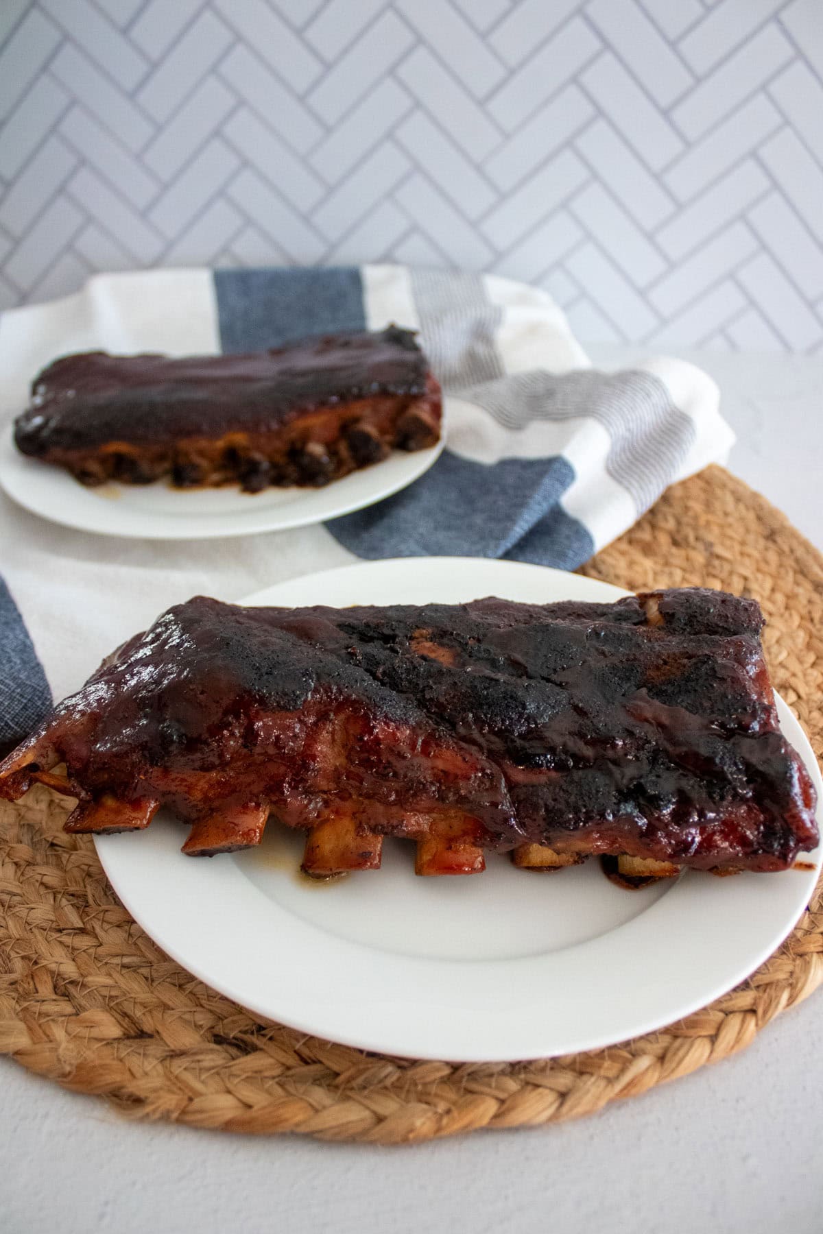 Two plates of glazed BBQ ribs are displayed on a table; one plate is in the foreground on a woven mat, while the other is in the background on a striped cloth, against a white chevron-patterned wall.