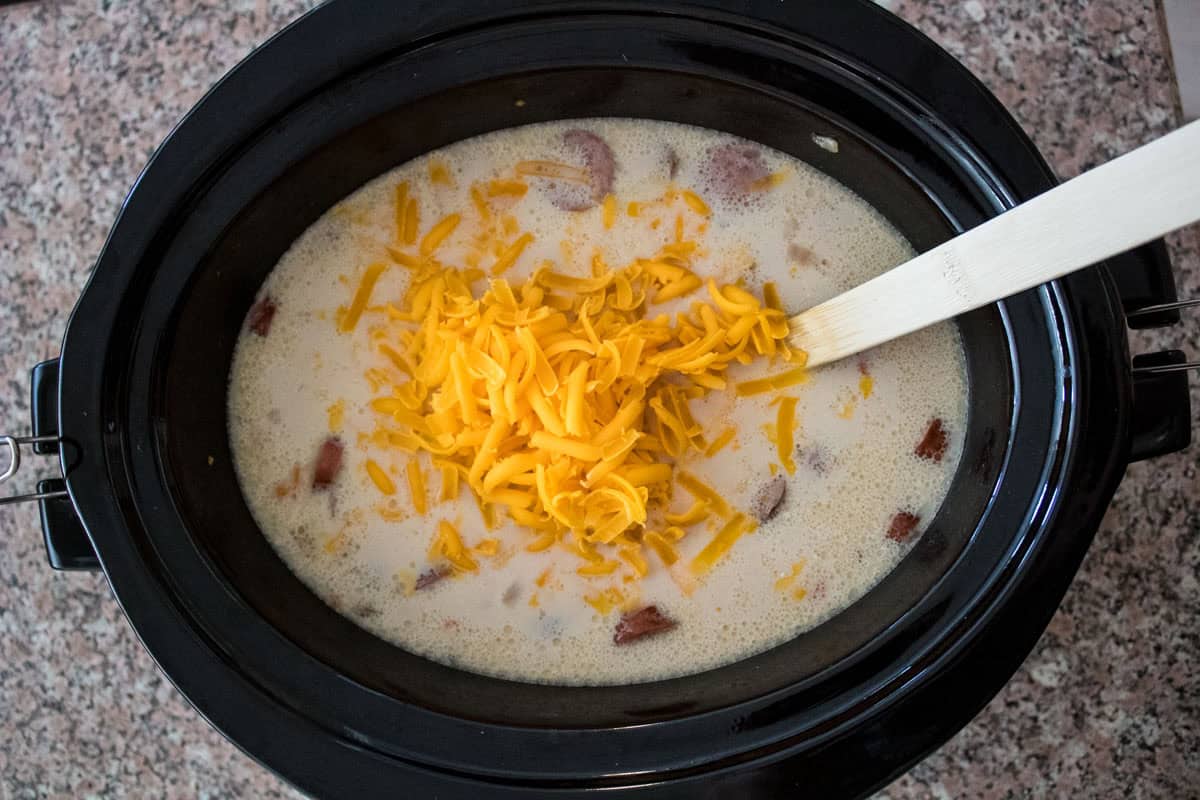 A slow cooker filled with a creamy soup, visible chunks of sausage, and topped with shredded cheddar cheese, with a wooden spoon resting inside.
