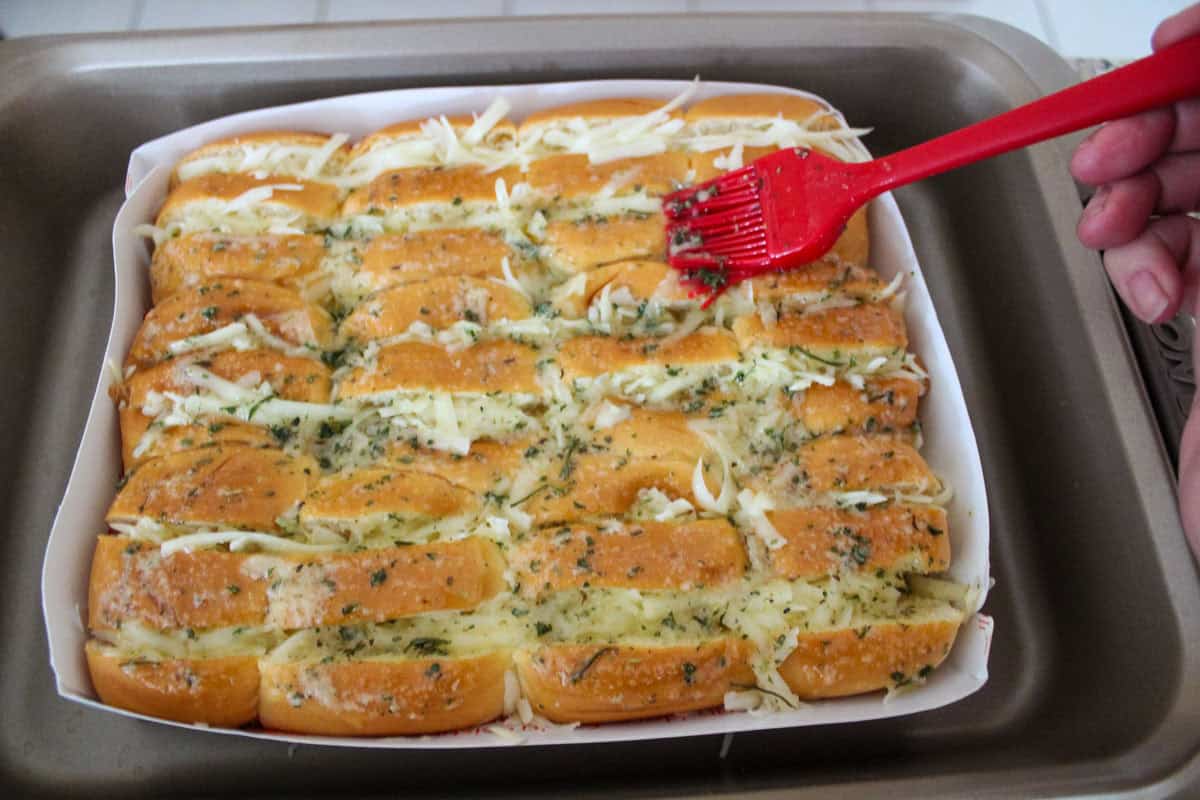 A hand uses a red brush to spread melted butter and herbs onto a batch of garlic bread rolls topped with shredded cheese, placed in a baking tray.