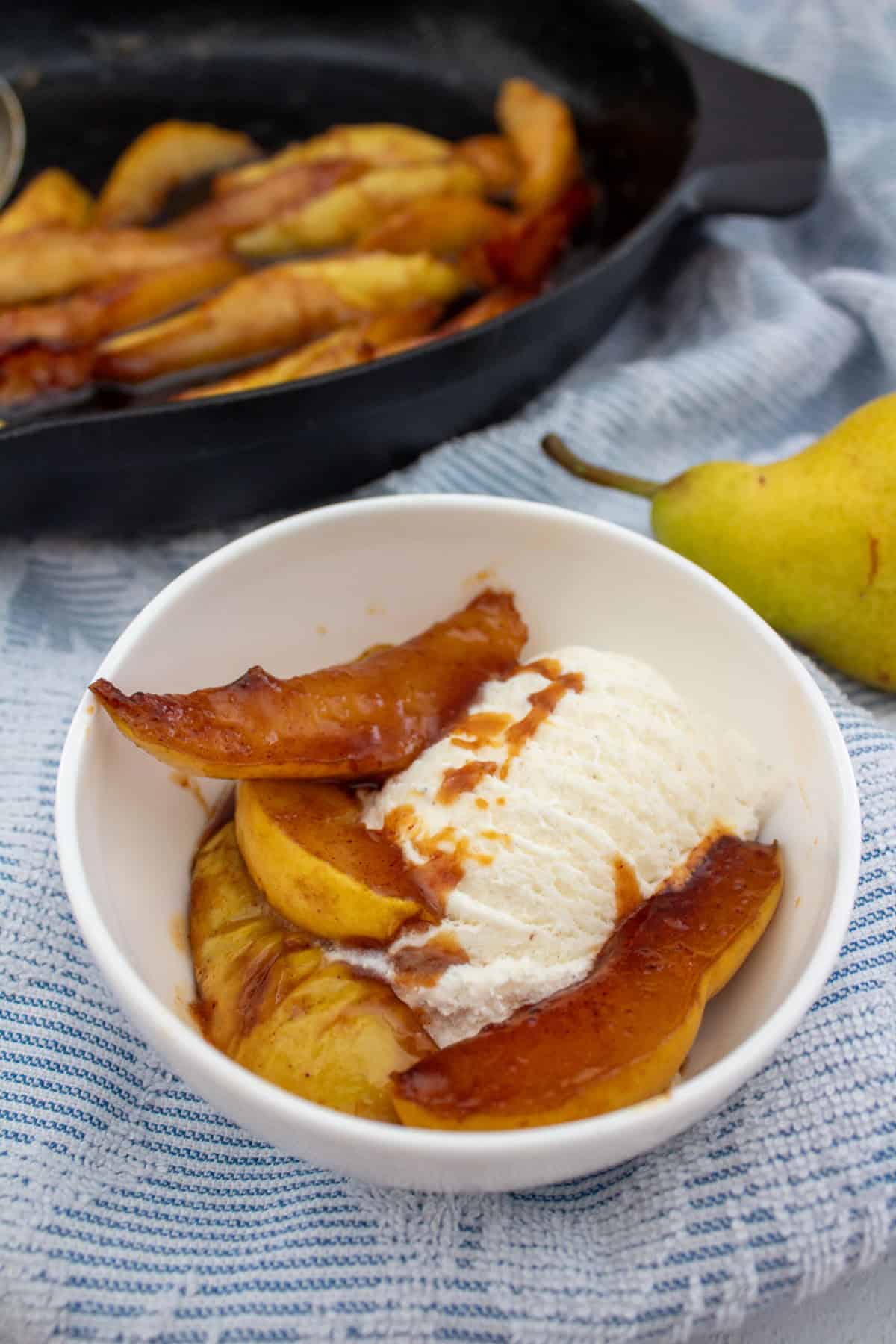 A white bowl with caramelized pear slices and a scoop of vanilla ice cream sits on a blue patterned cloth. A cooked pear dish in a black skillet and a fresh pear are in the background.