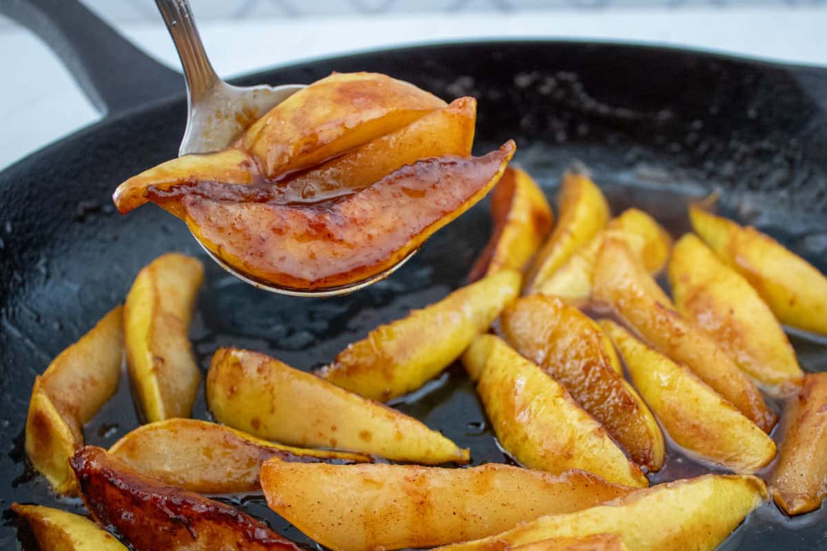 A close-up of caramelized apple slices being lifted from a skillet with a fork. The apples are golden brown and coated in a glossy sauce, with more slices visible cooking in the pan.