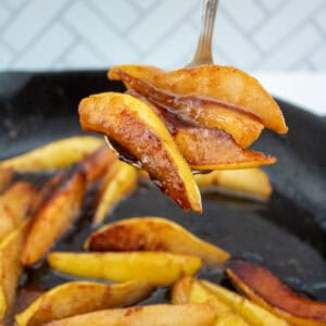 Sliced cooked pears with a golden brown caramelized coating, held up on a serving spoon above a skillet with more caramelized pear slices inside. The background features a white tile pattern.