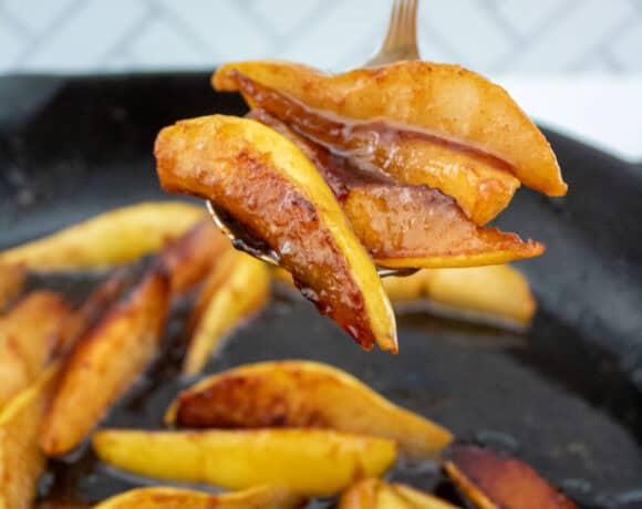 Sliced cooked pears with a golden brown caramelized coating, held up on a serving spoon above a skillet with more caramelized pear slices inside. The background features a white tile pattern.