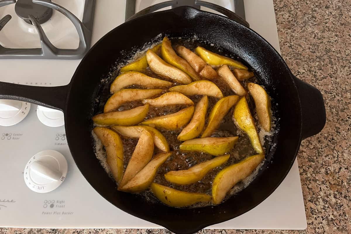 Sliced pears cooking in a cast iron skillet with bubbling liquid, likely sugar and spices, on a stovetop next to white burner knobs and a speckled countertop.