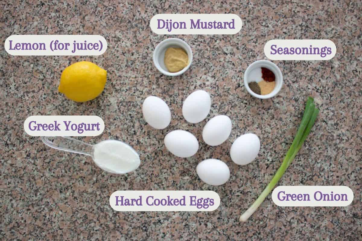 Ingredients for egg salad, including a lemon, cup of Greek yogurt, five hard-cooked eggs, Dijon mustard, assorted seasonings in a small dish, and a green onion, are arranged on a speckled countertop.