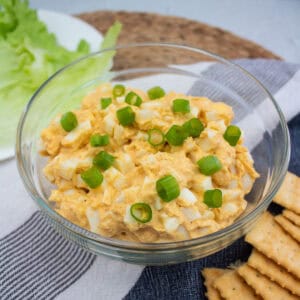 A clear glass bowl filled with creamy egg salad topped with chopped green onions, placed on a striped cloth next to crackers and leafy lettuce.