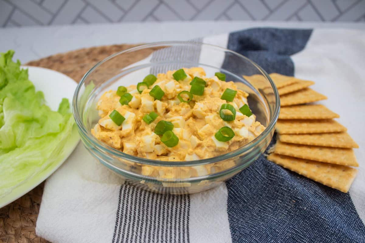 A glass bowl of egg salad topped with sliced green onions, placed on a striped cloth beside neatly arranged crackers and a plate of lettuce leaves.