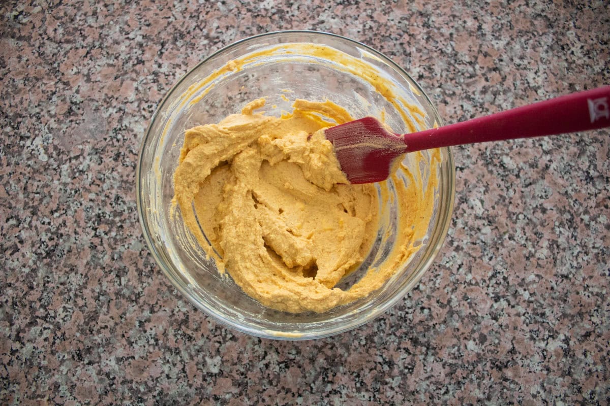 A glass bowl with thick, yellowish egg yolk mix being mixed by a red spatula, placed on a speckled countertop.