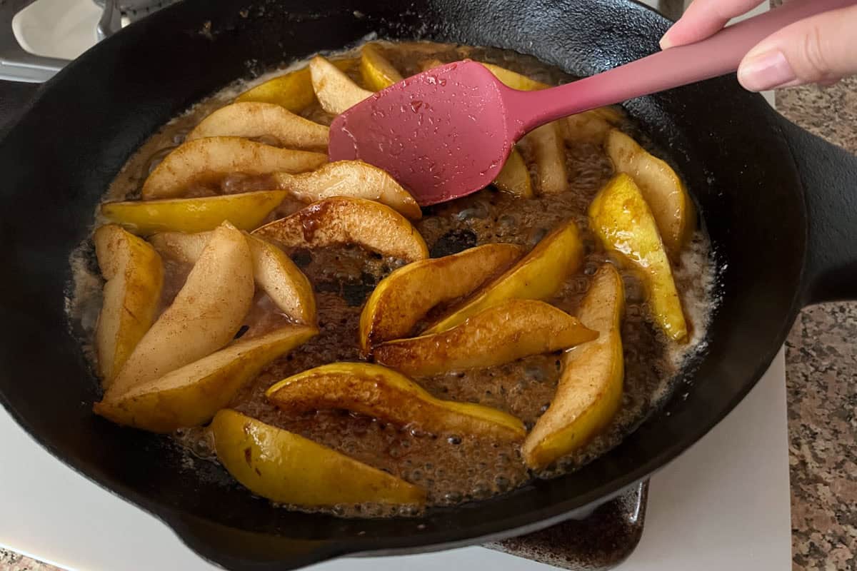 Sliced pears are being sautéed in a cast iron skillet with bubbling butter and cinnamon, while a hand flips them with a red spatula.