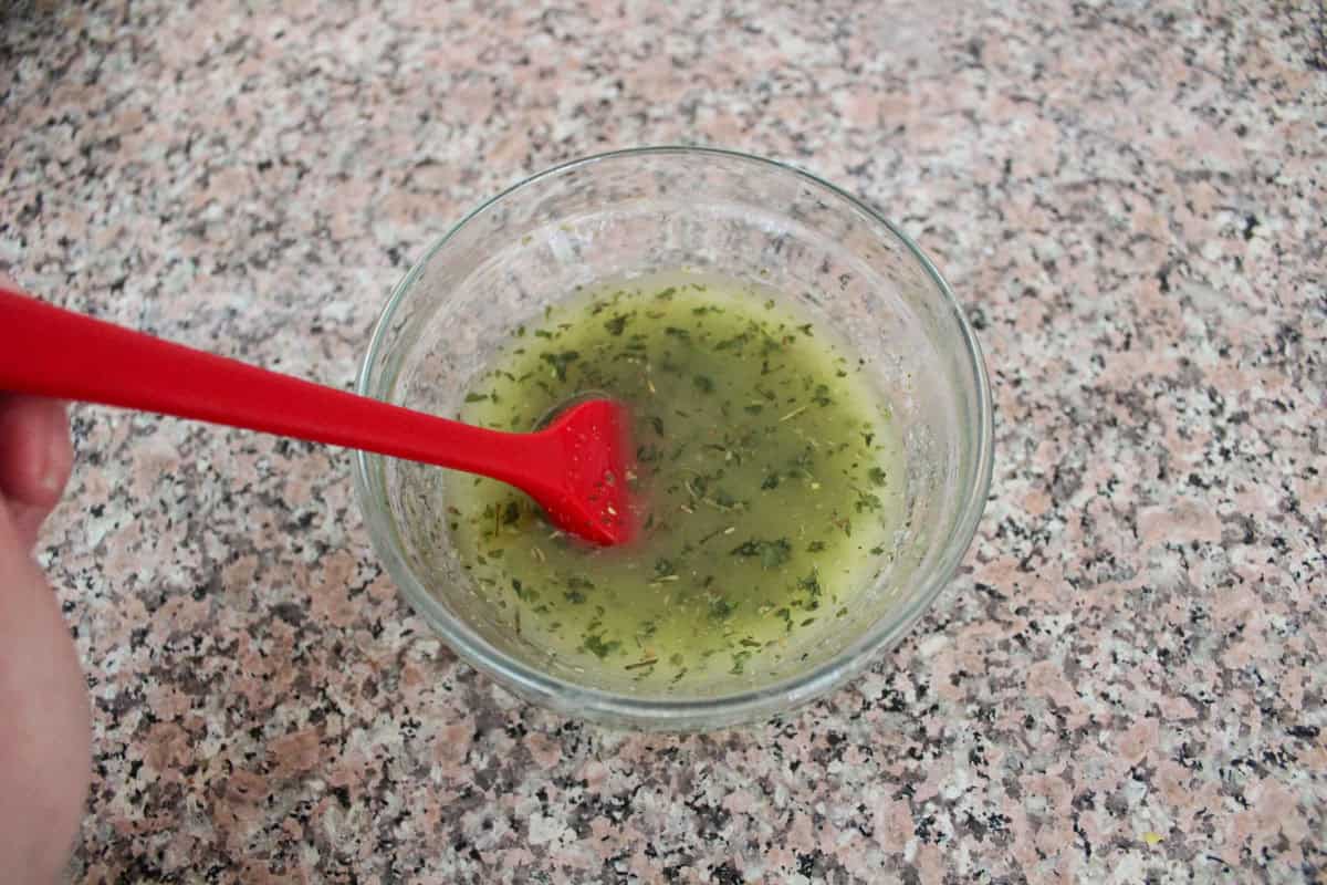 A hand holds a red spatula stirring a glass bowl filled with a greenish liquid mixture and herbs on a speckled countertop.