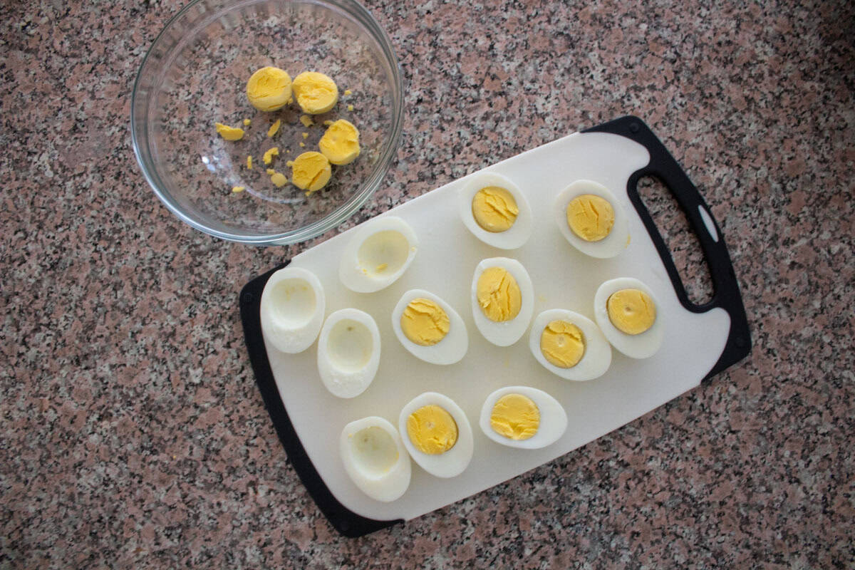 Halved hard-boiled eggs with yolks removed are arranged on a white cutting board. A glass bowl containing separated yolks and yolk pieces sits nearby on a speckled countertop.