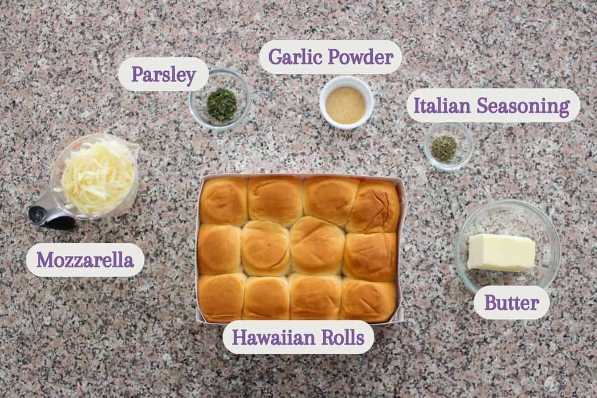 A top-down view of ingredients on a counter, labeled: parsley, garlic powder, Italian seasoning, shredded mozzarella, a tray of Hawaiian rolls, and a dish of butter.