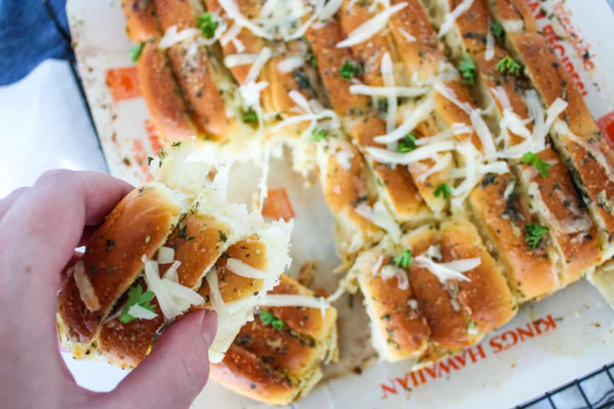 A hand holding a piece of cheesy, herb-topped pull-apart bread, with more pieces of the bread resting on a baking sheet in the background. The bread is garnished with shredded cheese and chopped parsley.
