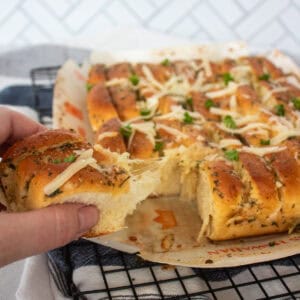 A hand pulls apart a piece of cheesy, herb-topped garlic bread from a baking sheet, with melted cheese stretching between the pieces and fresh parsley sprinkled on top.