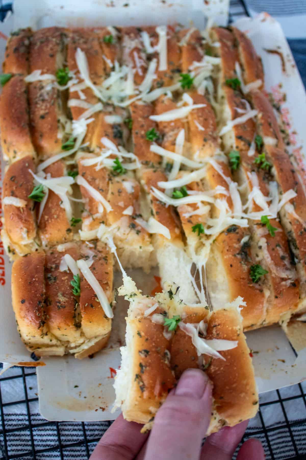 A hand holds a piece of soft, pull-apart garlic bread topped with shredded cheese and herbs, with the rest of the loaf on parchment tray in the background.