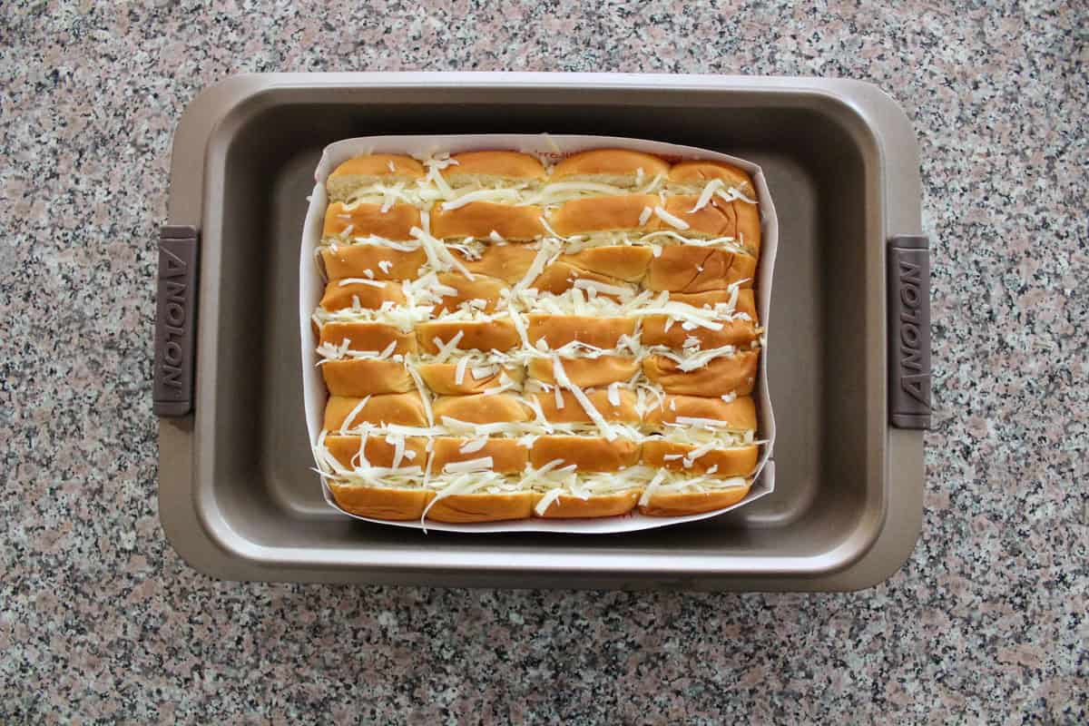 A baking tray on a granite countertop holds a batch of neatly arranged bread rolls stuffed with shredded cheese, placed in a baking tray.