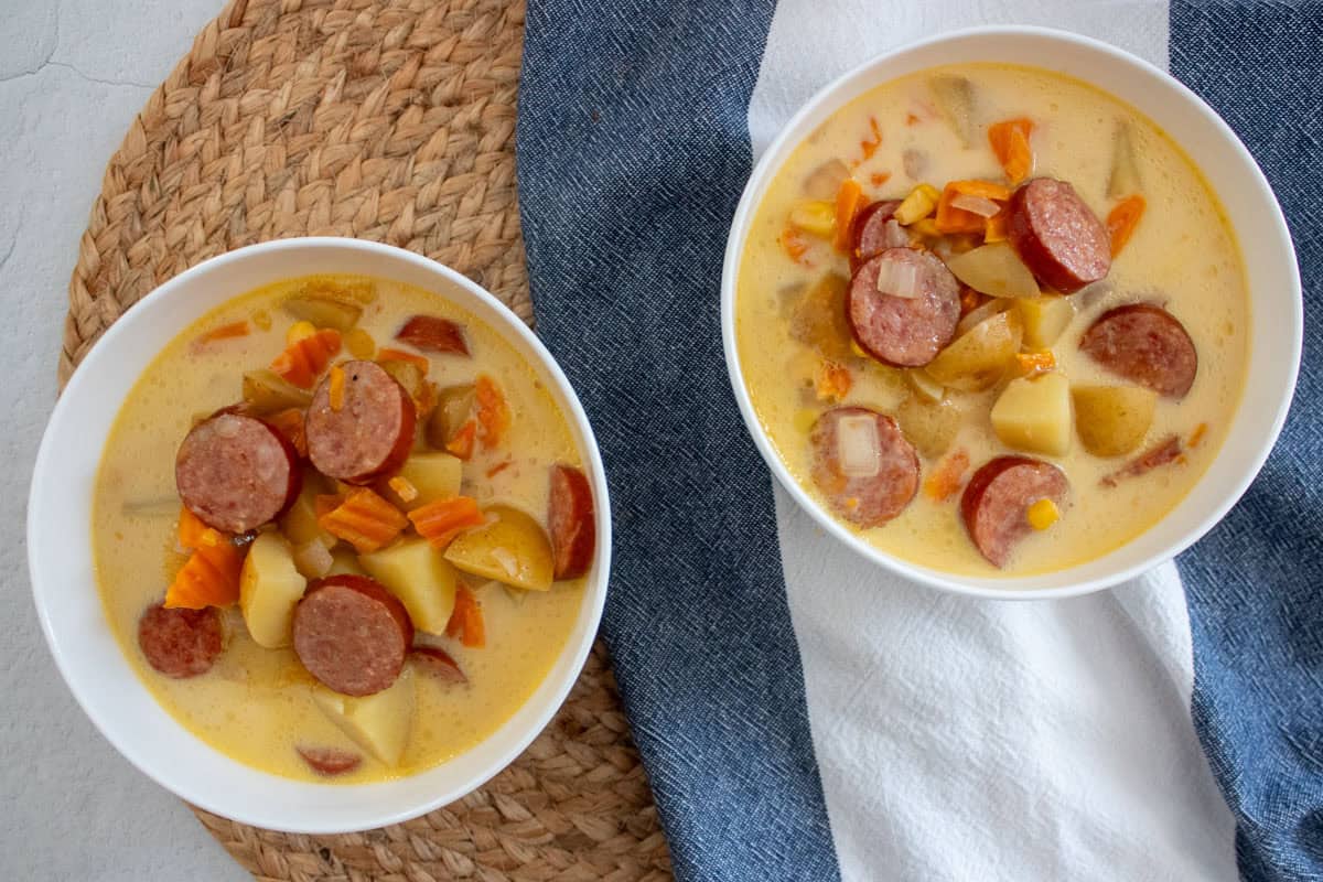 Two white bowls of creamy soup with sliced sausage, potatoes, and carrots on a white and blue cloth, placed on a woven placemat.