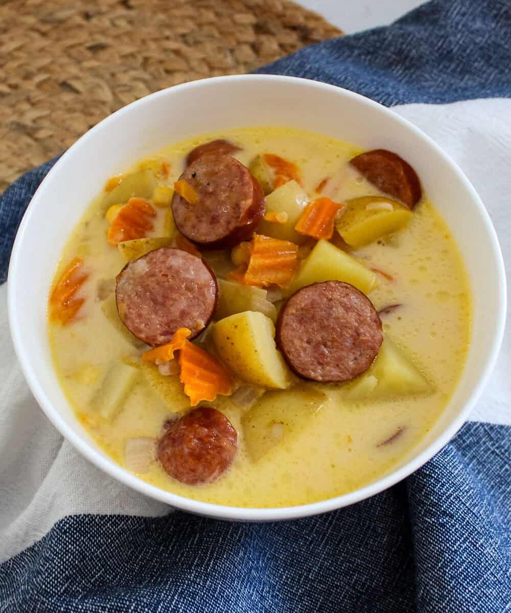 A white bowl of creamy soup containing sliced sausage, potatoes, and carrot pieces, set on a blue and white cloth with a woven placemat in the background.