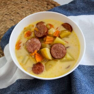 A white bowl of creamy soup containing sliced sausage, potatoes, and carrot pieces, set on a blue and white cloth with a woven placemat in the background.