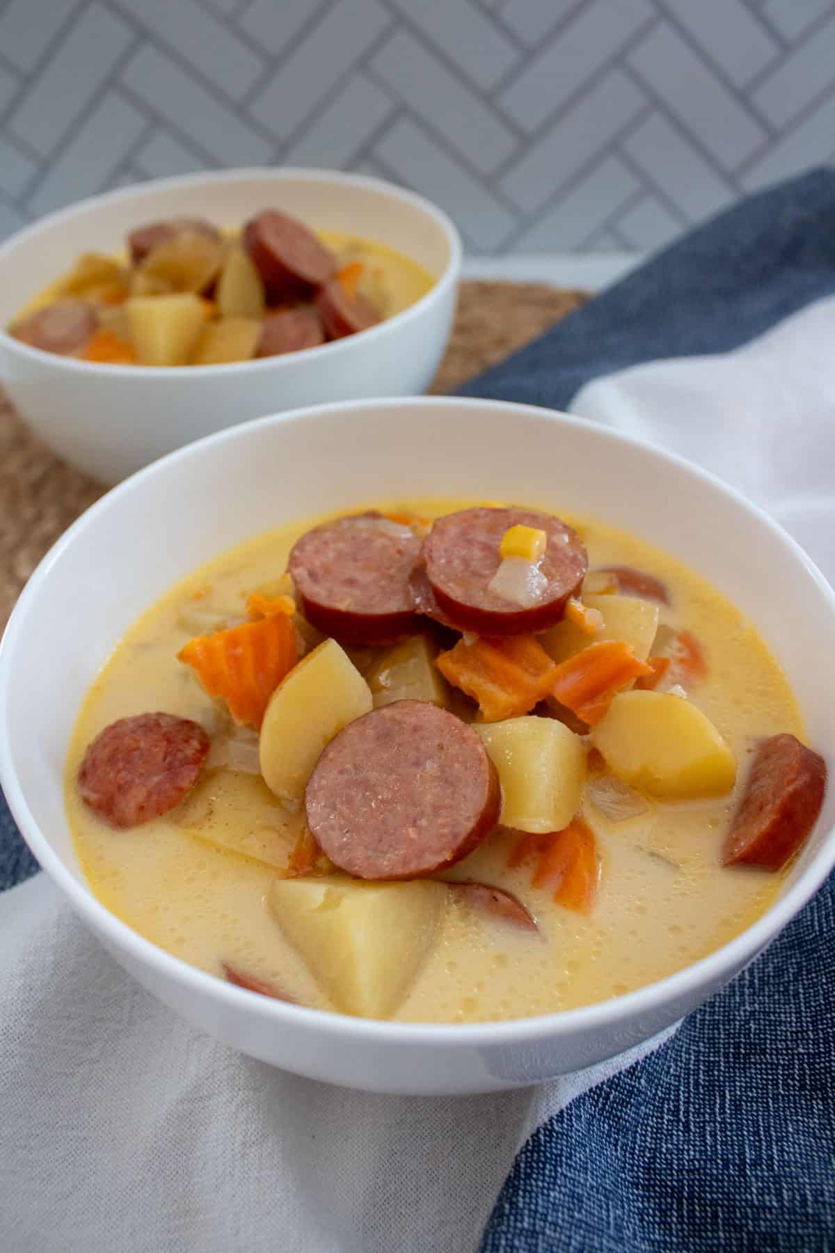 A white bowl filled with creamy soup containing sliced sausage, potatoes, carrots, and corn, with another similar bowl in the background. The scene includes a white napkin and a woven placemat.