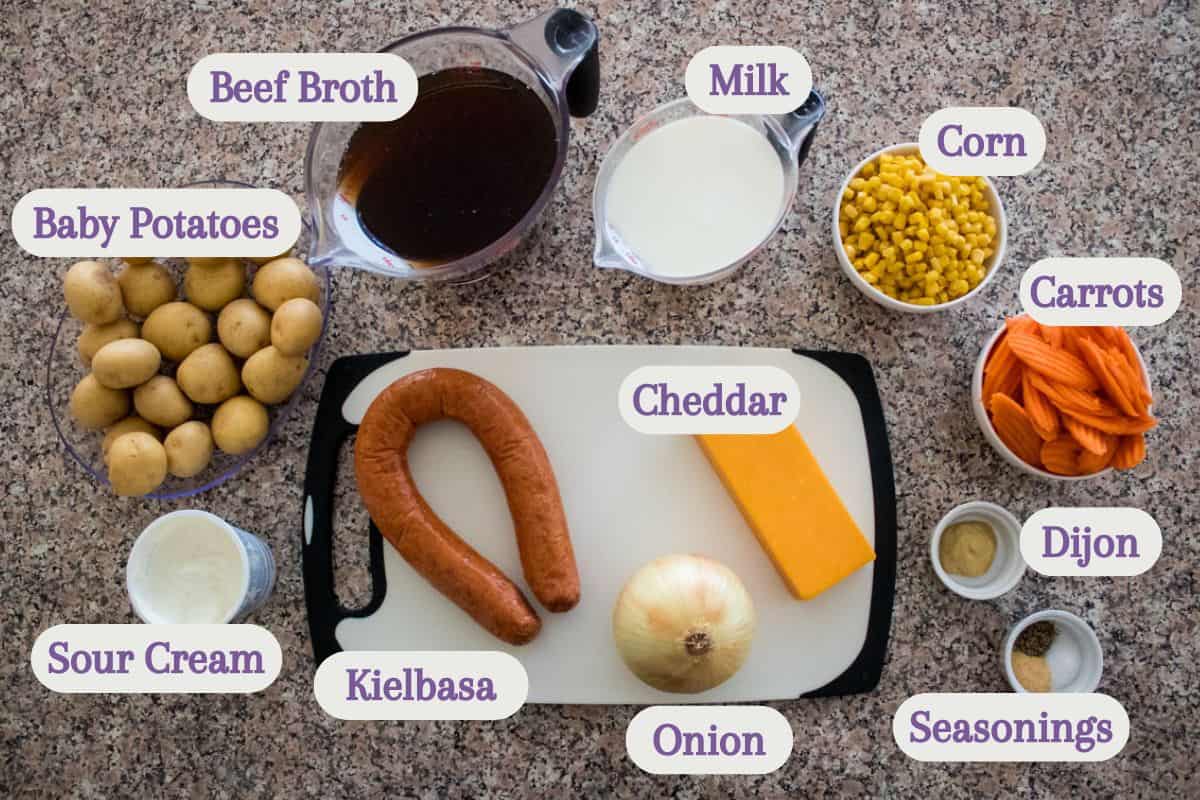 Ingredients for a recipe are arranged on a countertop, each in a separate bowl or container, and labeled: beef broth, milk, corn, carrots, seasonings, dijon, cheddar, onion, kielbasa, sour cream, and baby potatoes.