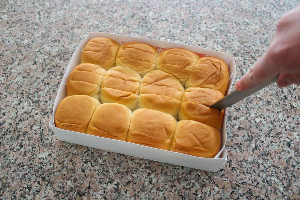 A hand is using a knife to cut into a tray of soft, golden-brown dinner rolls on a speckled countertop.