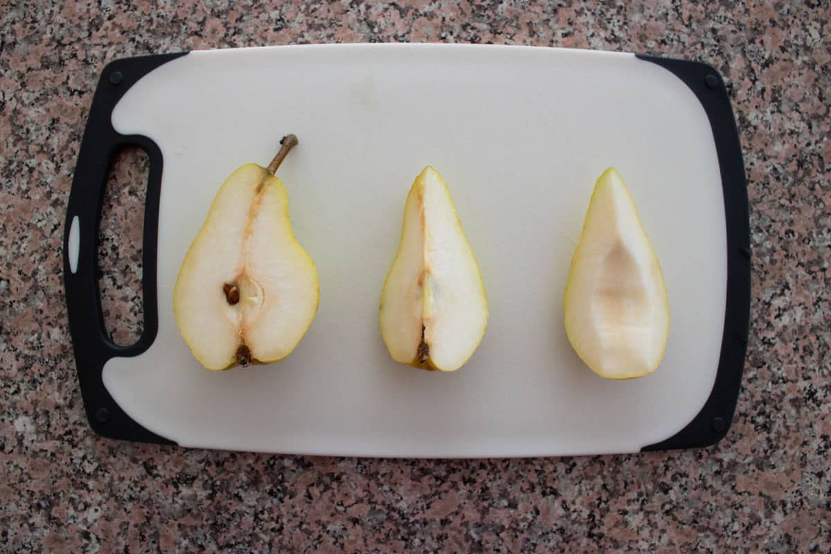 A pear cut into three sections—one half with seeds, one quarter with stem, and the last quarter hollowed-out—sits on a white cutting board on a speckled countertop.