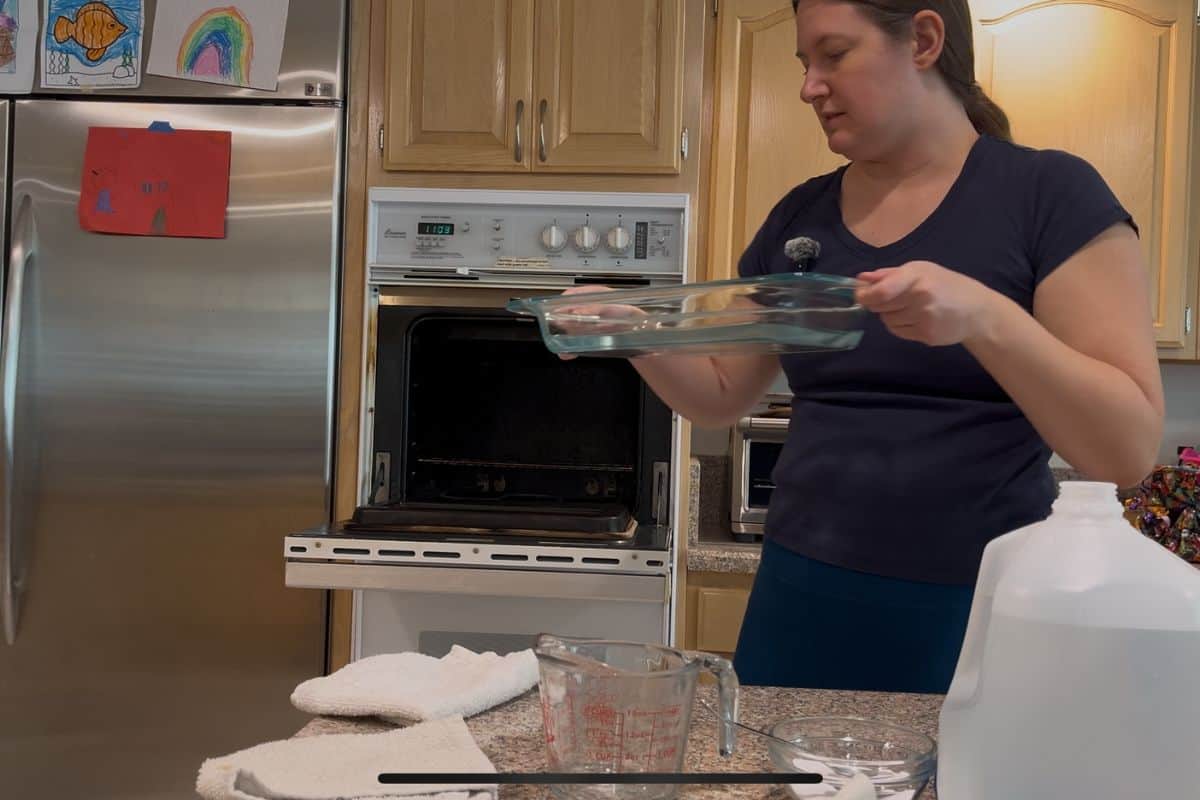A woman in a kitchen holds a glass baking dish near an open oven, ready to steam clean the oven. Towels, a measuring cup, and a jug of distilled water sit on the counter.