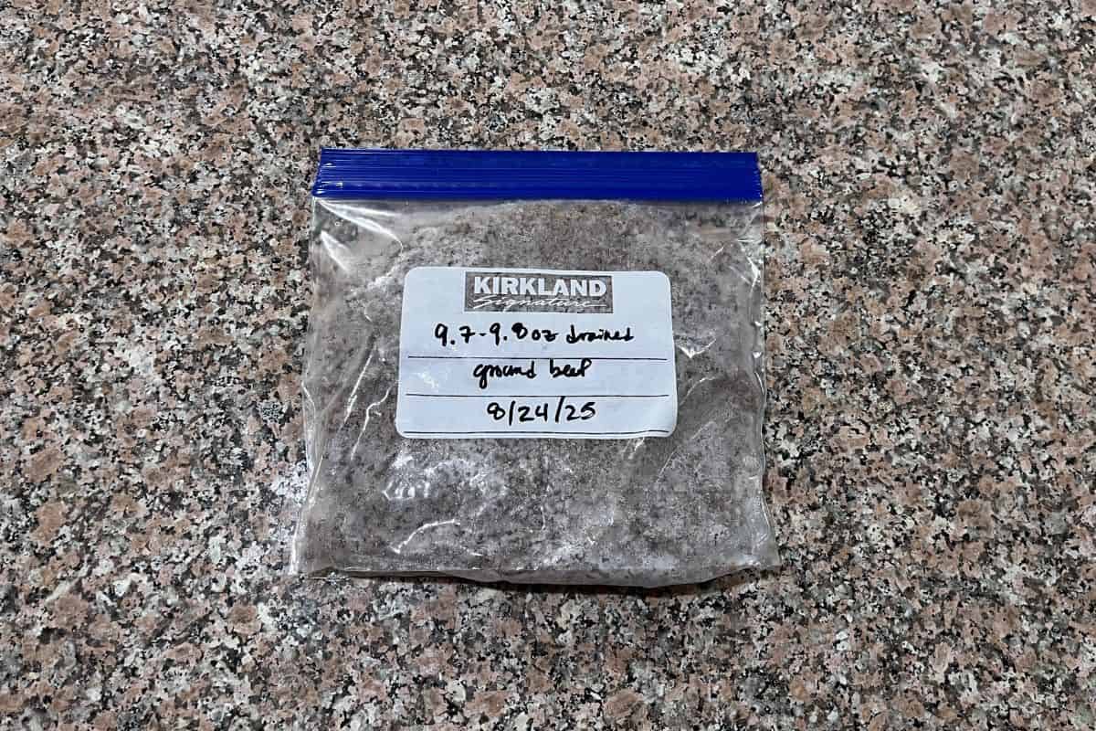 A clear plastic bag with a blue zip-top, labeled Kirkland and handwritten notes of the contents, date, and weight. The bag contains frozen ground beef and rests on a speckled countertop.