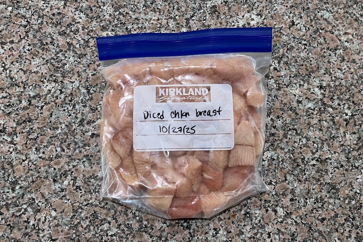 A ziplock bag labeled Diced chkn breast 10/27/25 contains diced raw chicken breast pieces. The bag is placed on a speckled granite countertop.