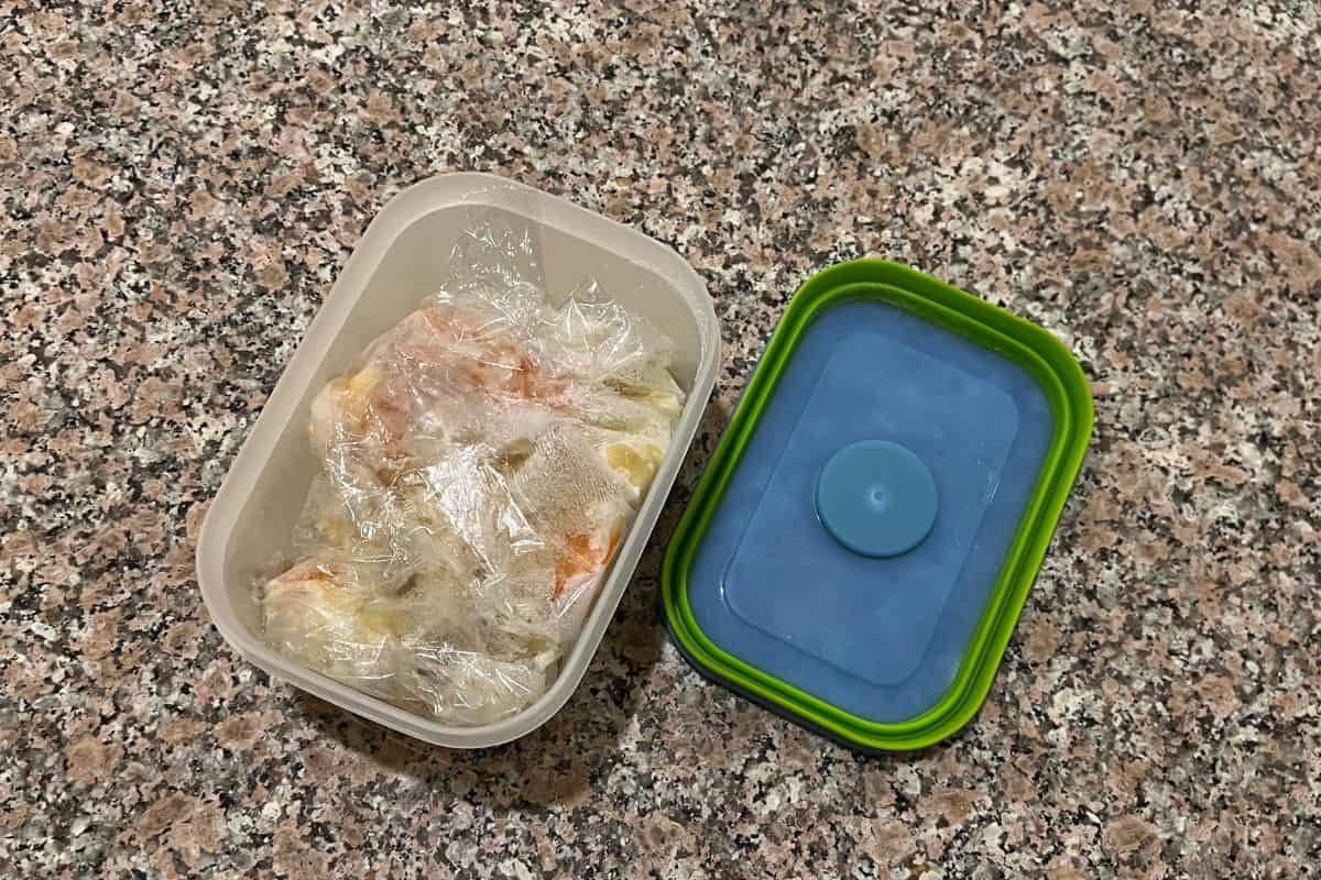 A plastic food container with a green-edged blue lid sits on a speckled countertop. The container is open, showing food wrapped in clear plastic wrap inside.