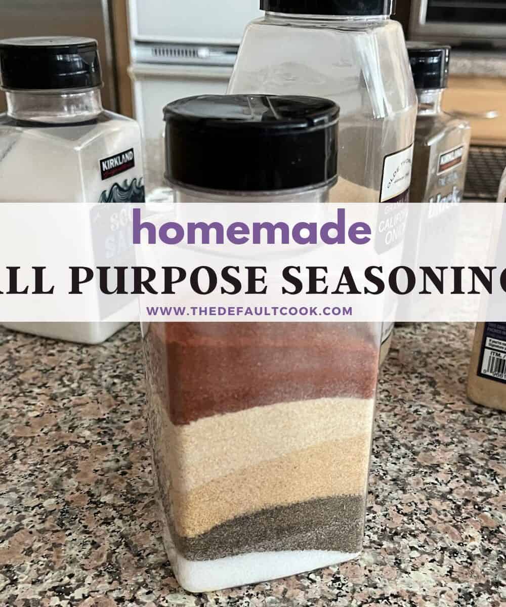 A clear container filled with layers of different spices sits on a kitchen countertop, surrounded by other spice containers. The label reads homemade ALL PURPOSE SEASONING www.thedefaultcook.com.