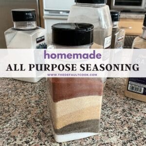 A clear container filled with layers of different spices sits on a kitchen countertop, surrounded by other spice containers. The label reads homemade ALL PURPOSE SEASONING www.thedefaultcook.com.