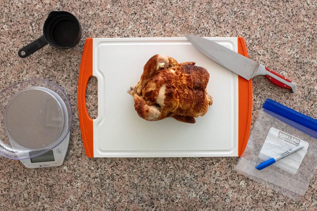 A whole cooked rotisserie chicken sits on a white cutting board with orange handles. Nearby are a large knife, a digital kitchen scale, a measuring cup, and a resealable plastic bag with a marker.