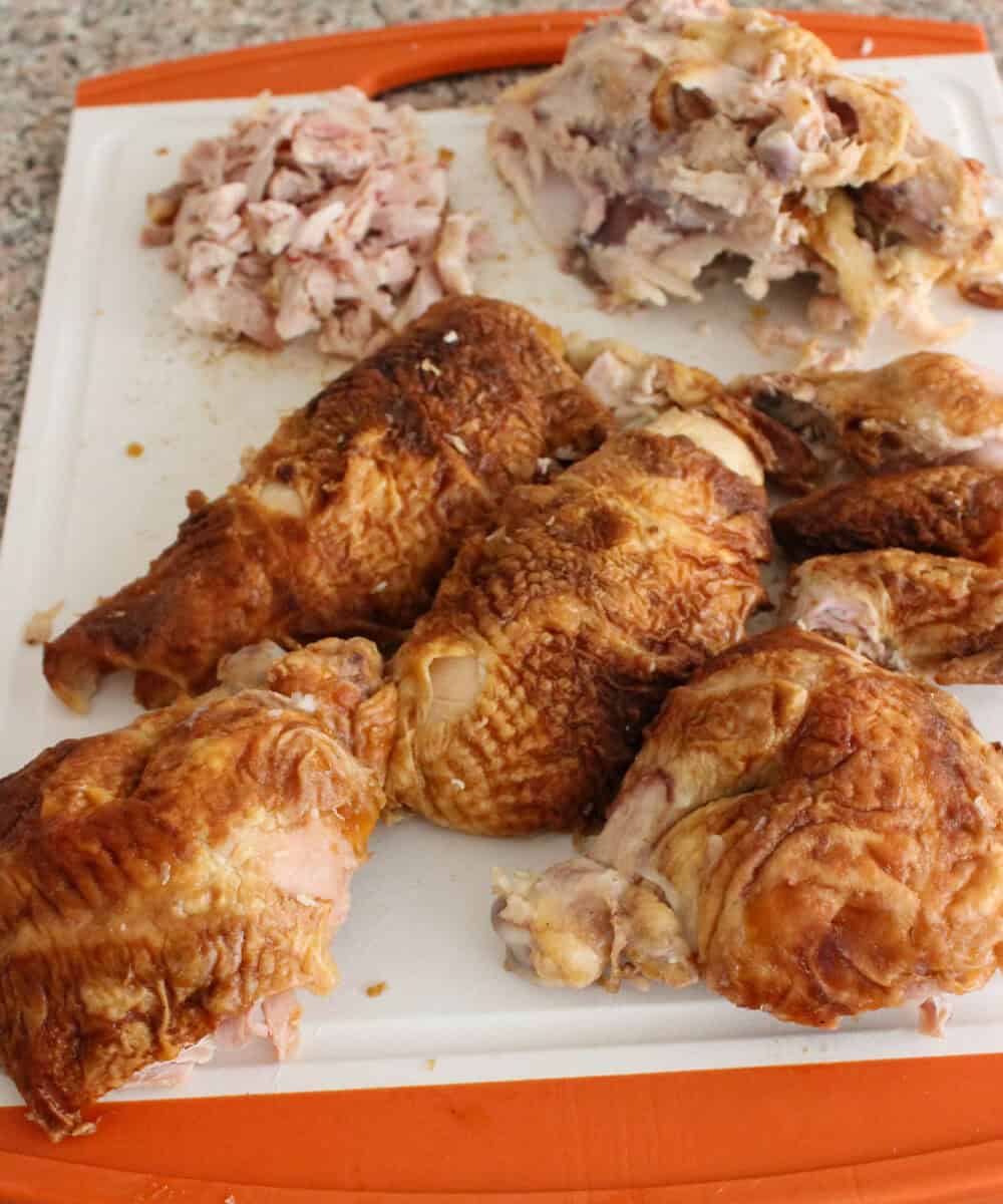 Various pieces of cooked, rotisserie chicken are arranged on a white and orange cutting board, with shredded and chopped chicken meat in the background. The cutting board is on a speckled countertop.