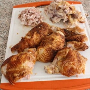 Various pieces of cooked, rotisserie chicken are arranged on a white and orange cutting board, with shredded and chopped chicken meat in the background. The cutting board is on a speckled countertop.