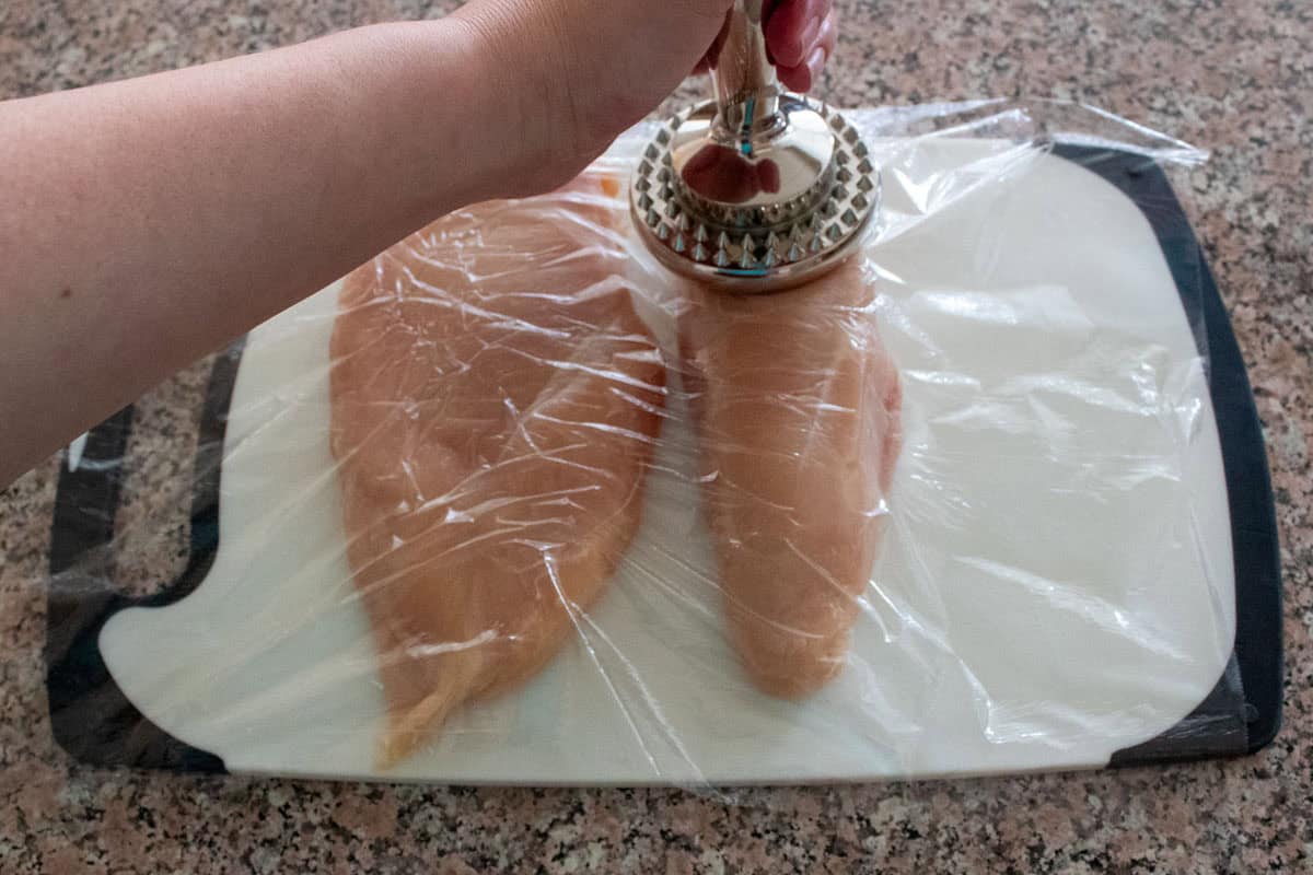 A hand uses a meat tenderizer to pound two raw chicken breasts covered with plastic wrap on a white cutting board, placed on a speckled countertop.